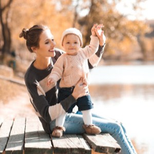 Smiling woman holding baby girl wearing knitted casual clothes sitting in autumn park. Happiness.