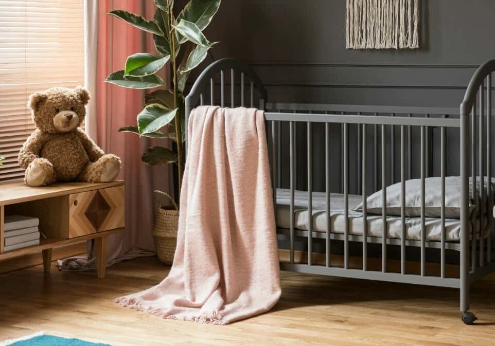 Real photo of a baby crib standing between a low cupboard and an armchair, lamp and stool in child's room interior with wooden floor and grey walls with molding