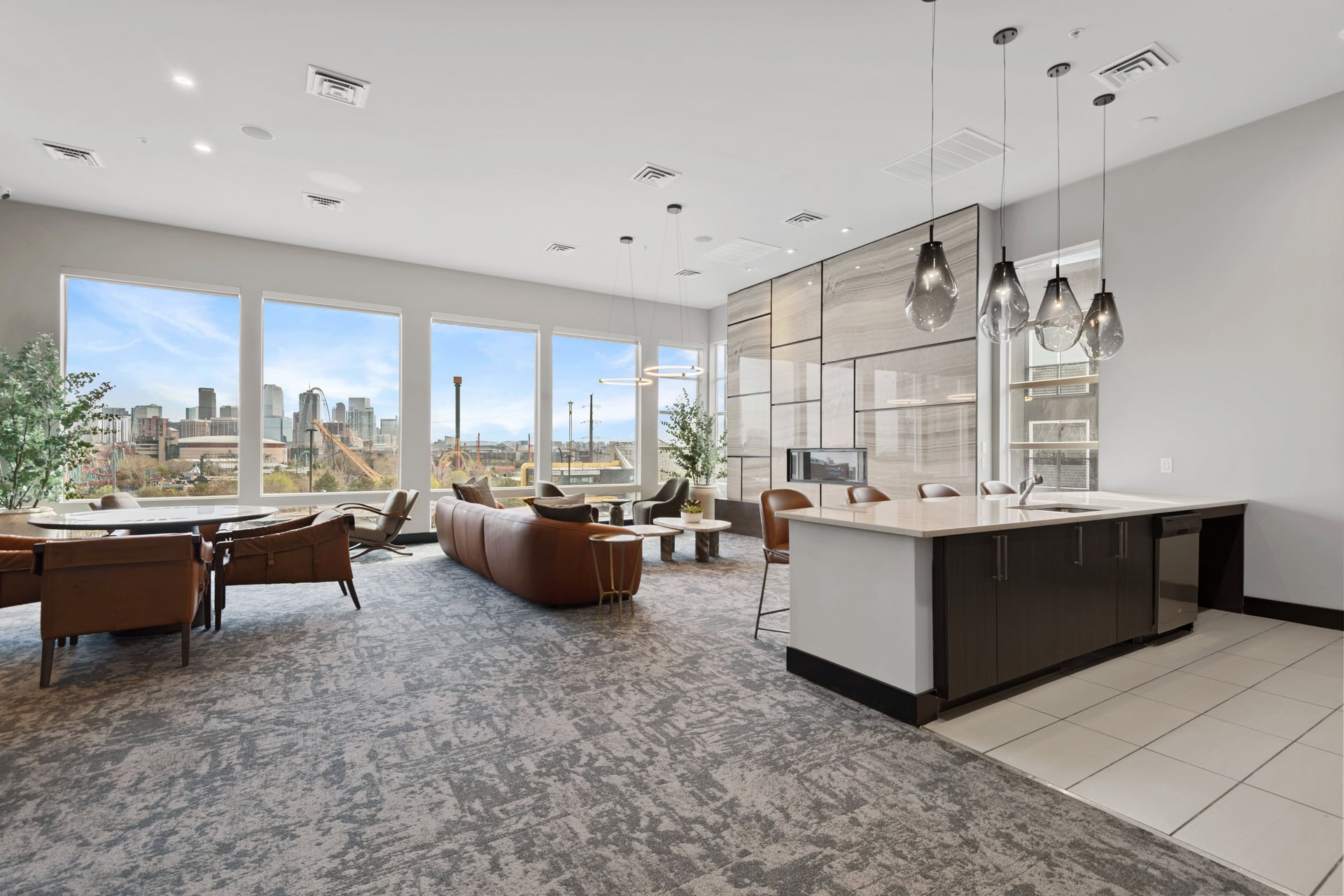 Modern open-concept lounge with large windows, city skyline views, Element 47 brown leather seating, round dining table, and a sleek kitchen island with pendant lights. Neutral tones and natural light create a welcoming atmosphere.