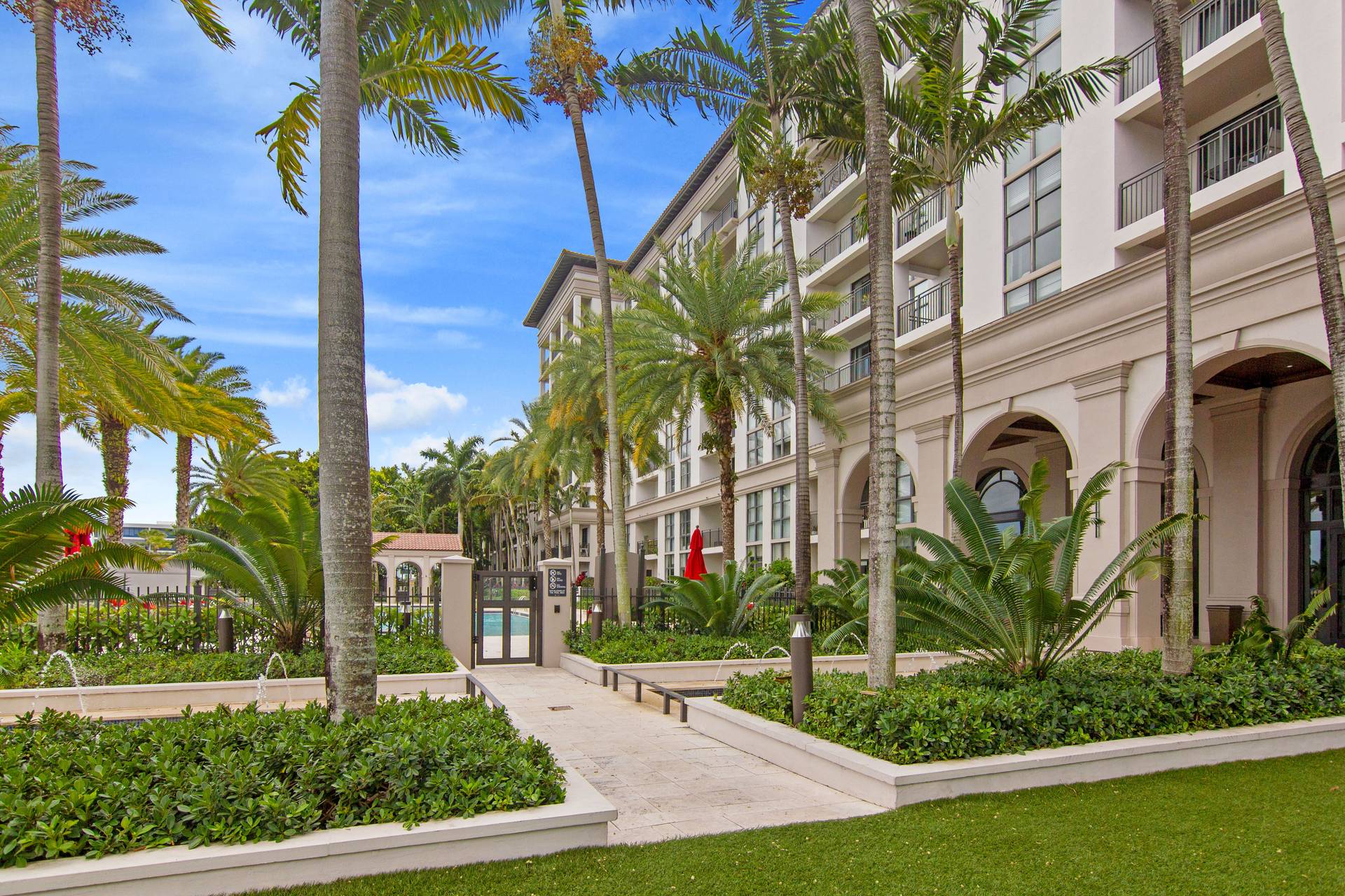 A luxurious hotel courtyard at Windsor at Doral features palm trees, lush green plants, a walking path, benches, and a pool area in the background beside a modern multi-story building under a blue sky.