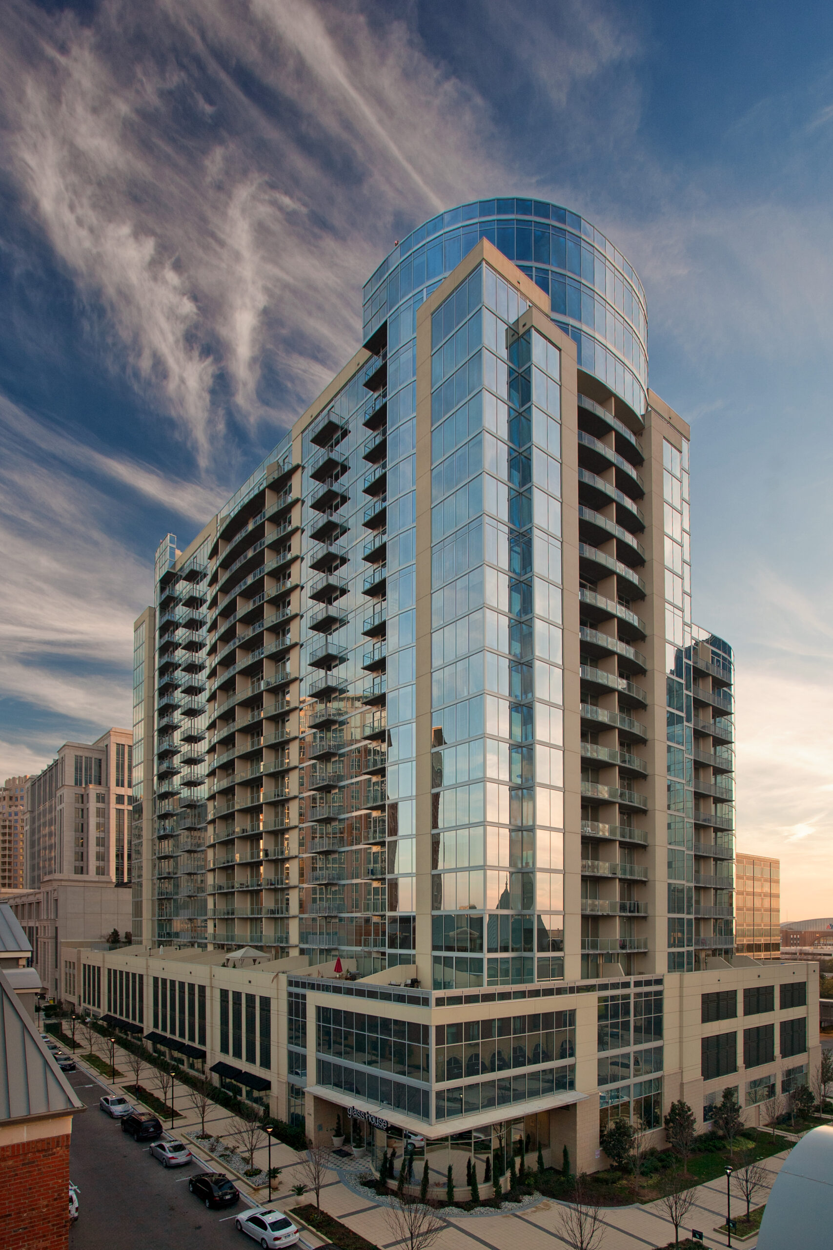 A modern high-rise apartment building, Glass House by Windsor, features many glass windows and balconies reflecting the sky, standing in an urban area with parked cars and a partially cloudy blue sky overhead.