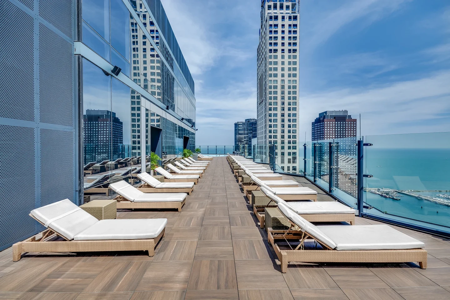 A rooftop deck with rows of white cushioned lounge chairs faces glass railings, inviting you to savor each moment while overlooking tall city buildings and a vast body of water beneath a blue sky.