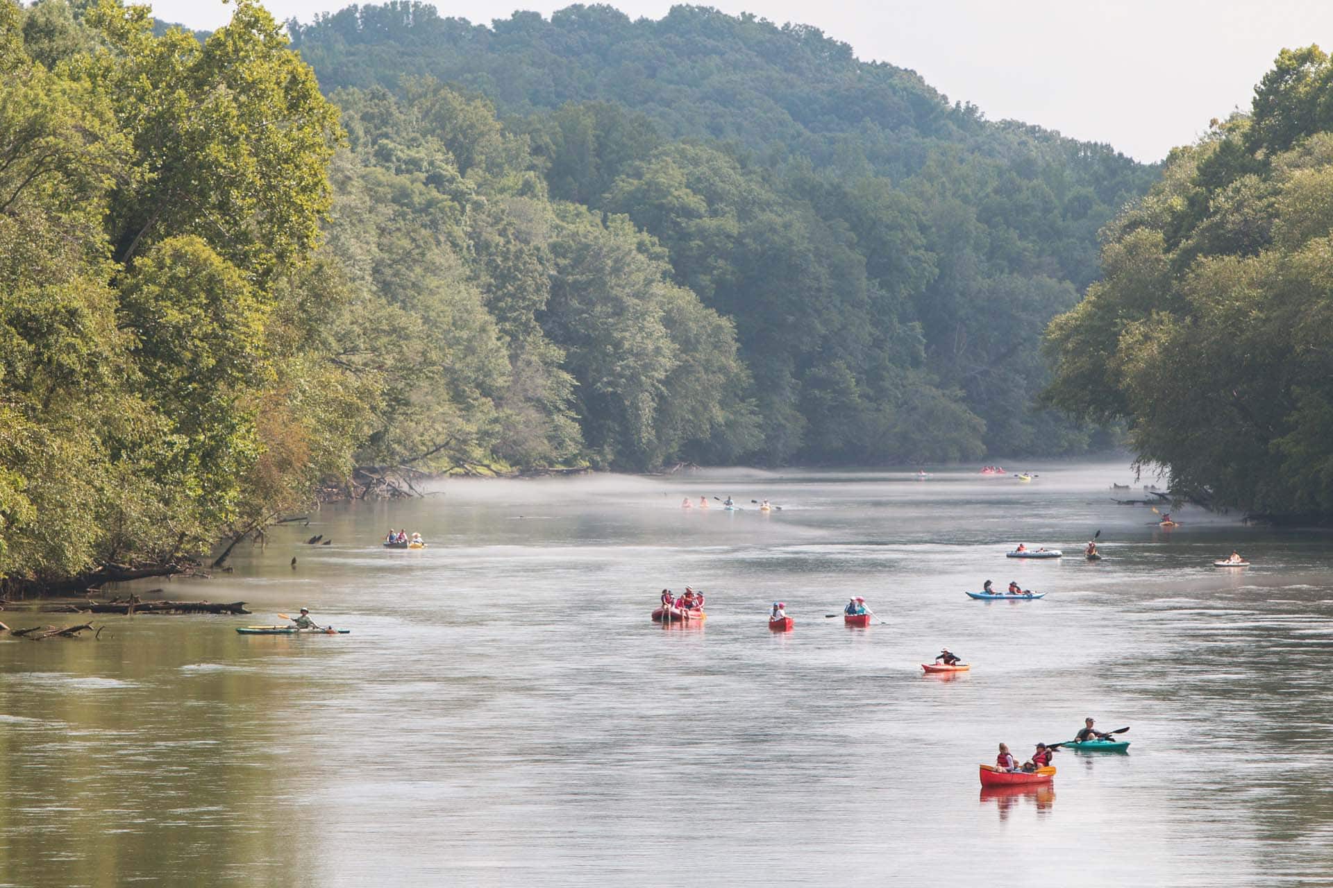 A river with several people kayaking and canoeing, surrounded by dense green trees and distant hills.