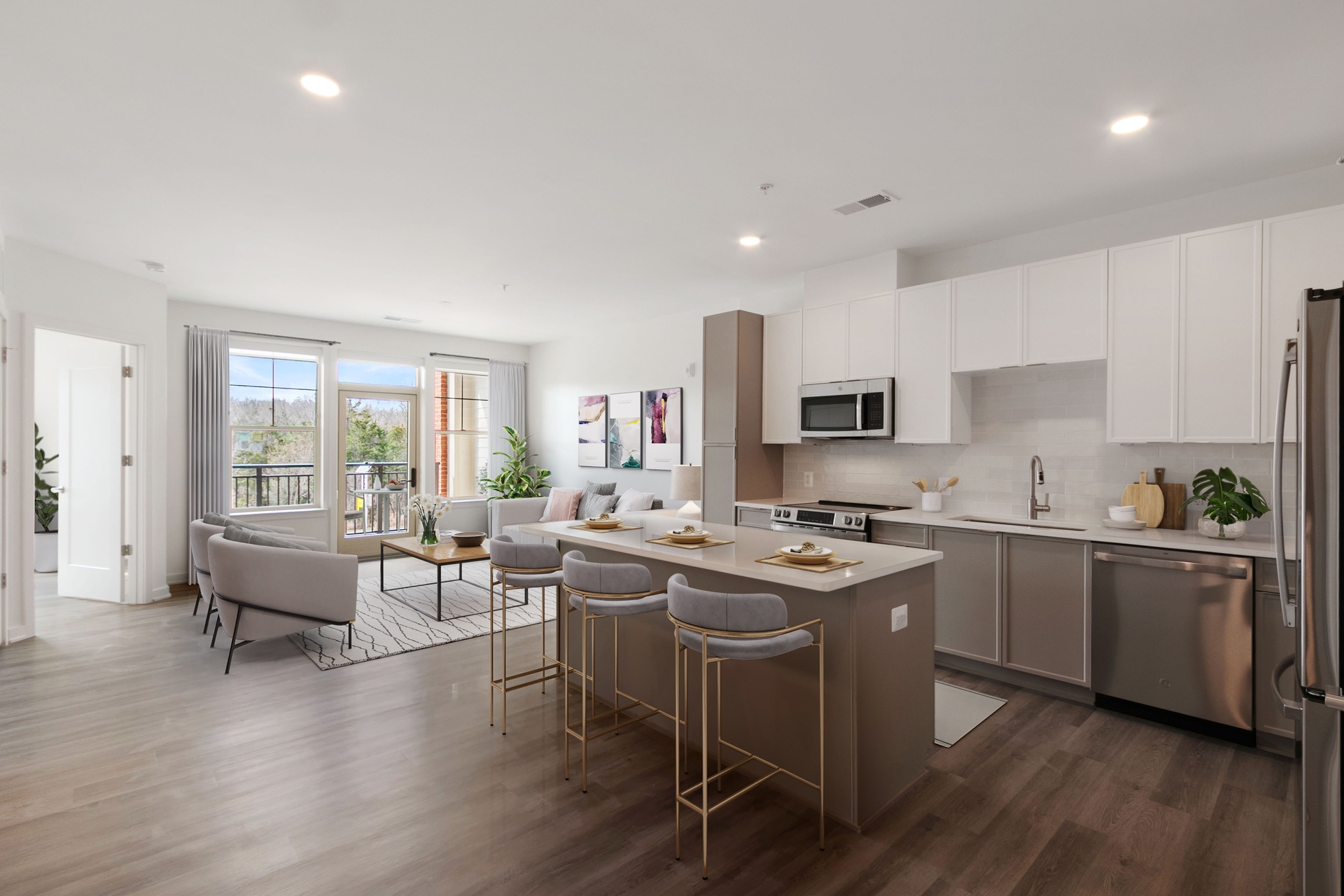 Modern open-concept kitchen and living room at Windsor Leesburg with a central island, three barstools, stainless steel appliances, white cabinets, a gray sofa, and large windows letting in natural light.