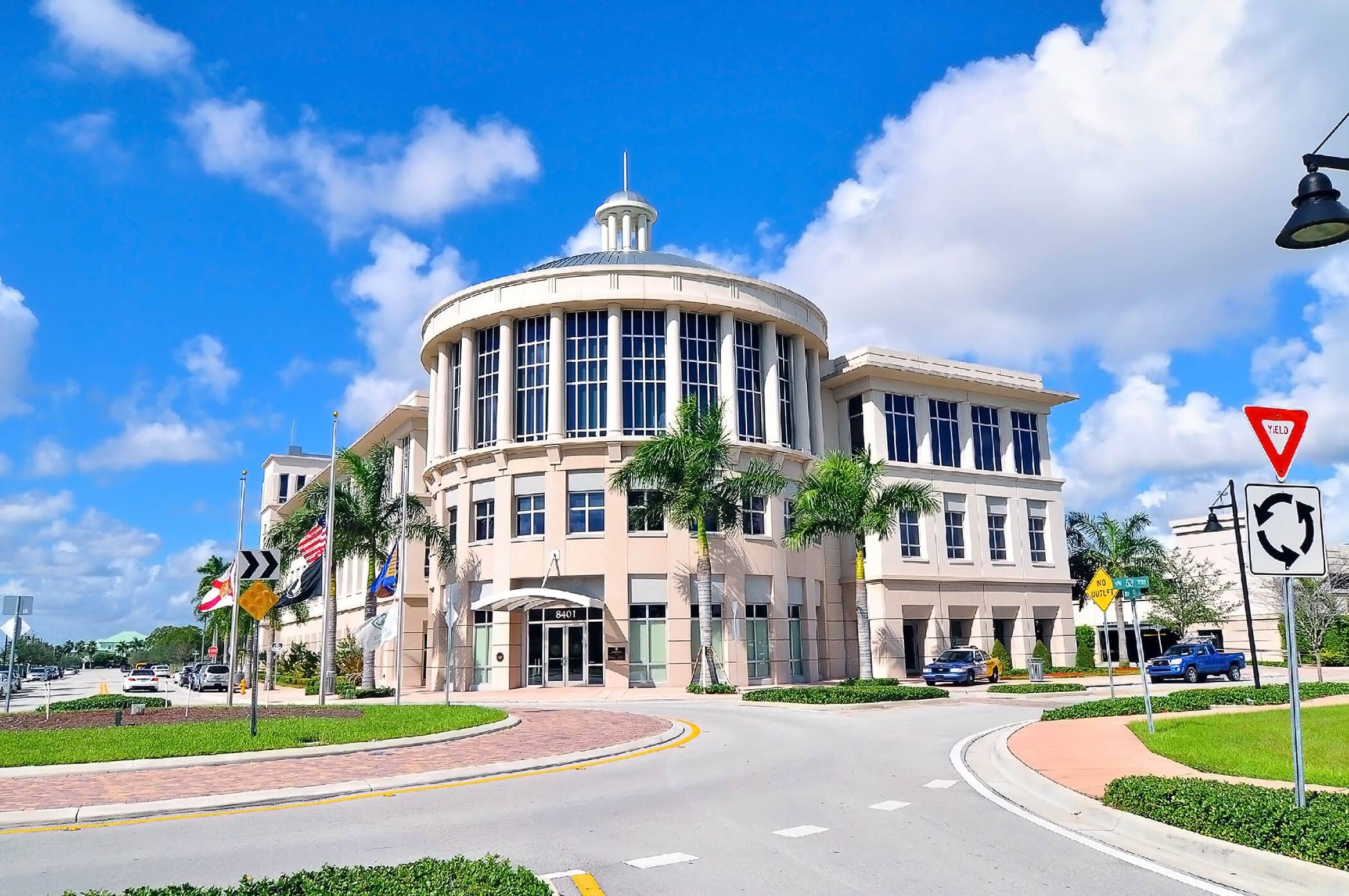 A modern, light-colored government building with tall windows and palm trees in front sits at a roundabout beneath a bright blue sky. Near the entrance, US and state flags wave—just steps from Centrico by Windsor.