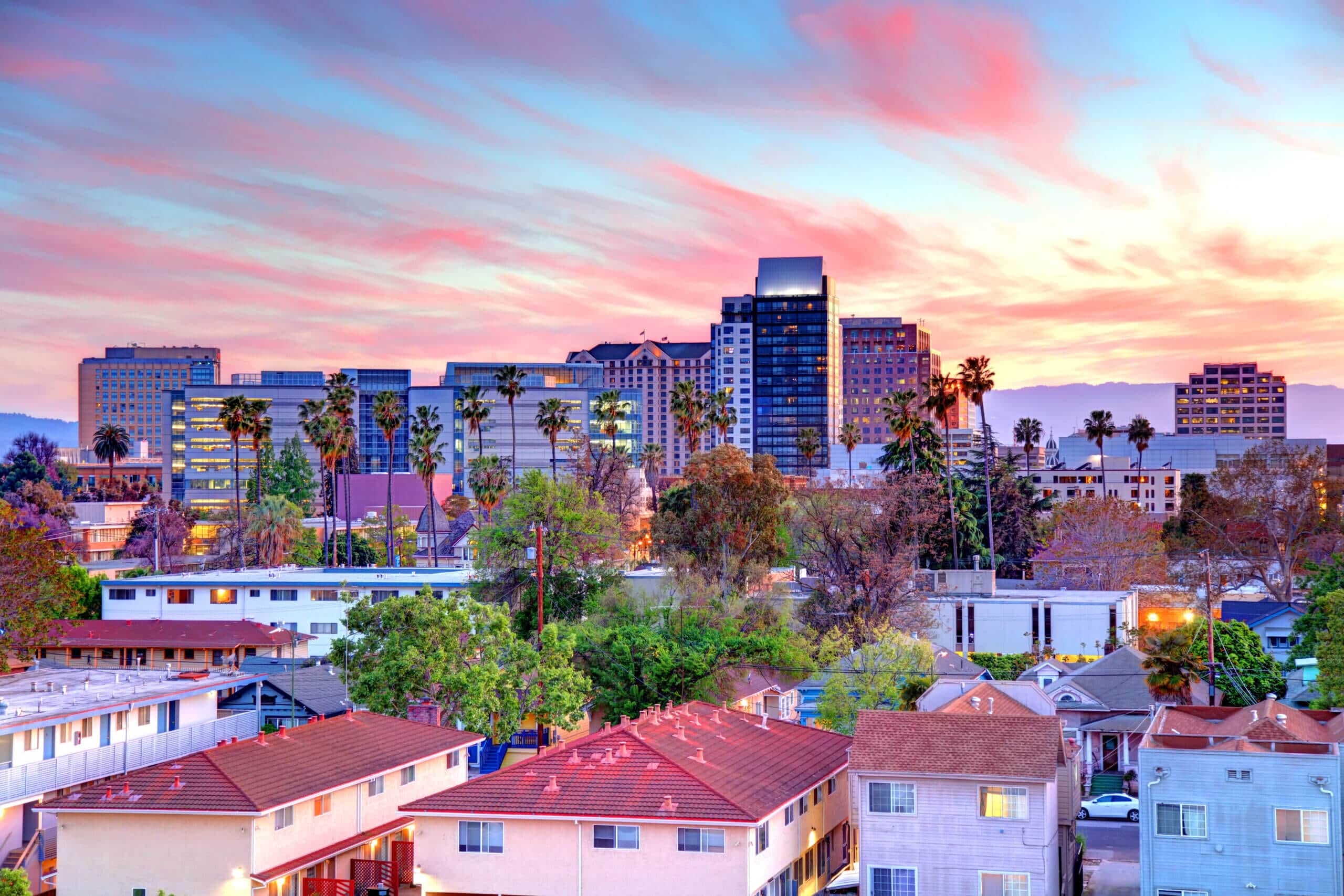 A colorful sunset sky over Cannery Park by Windsor cityscape with modern high-rise buildings, tall palm trees, and residential houses with red roofs in the foreground, creating a vibrant urban scene.