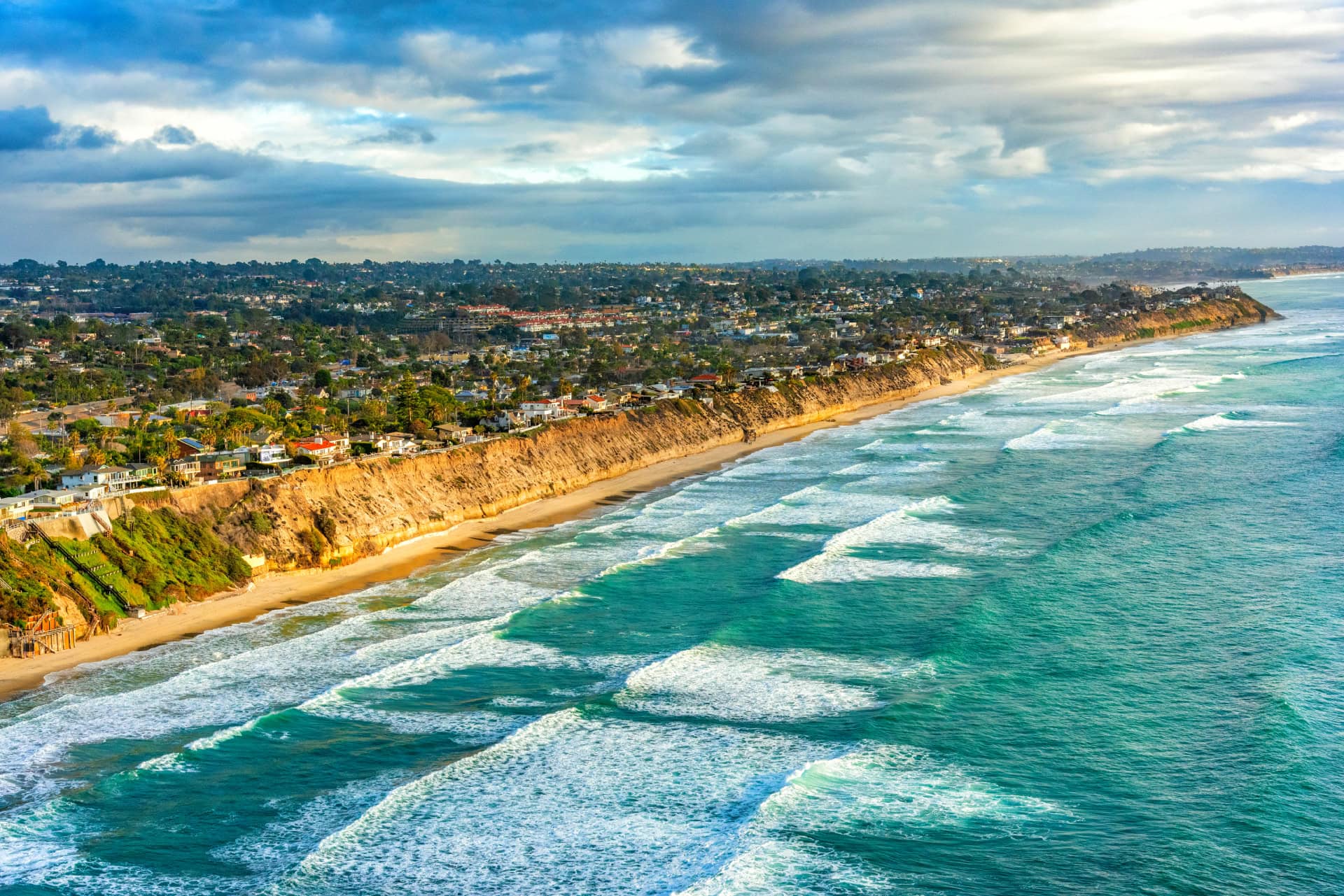 Aerial view of a coastal city with waves crashing onto a sandy beach lined with cliffs, houses, and greenery under a partly cloudy sky.