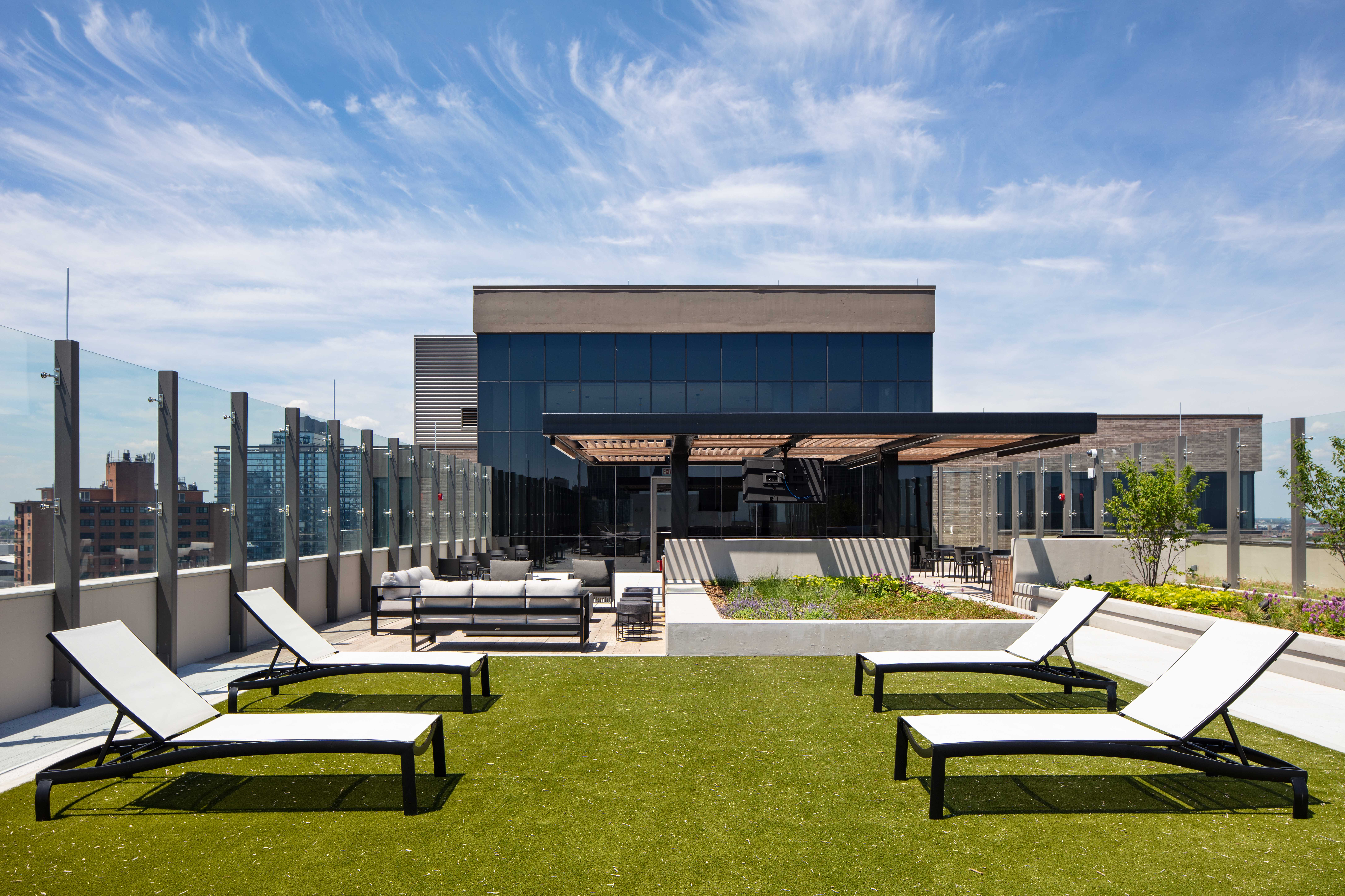 Modern rooftop terrace with lounge chairs on artificial grass, outdoor seating area under a canopy, glass railings, planter boxes, and city buildings in the background under a blue sky with wispy clouds.