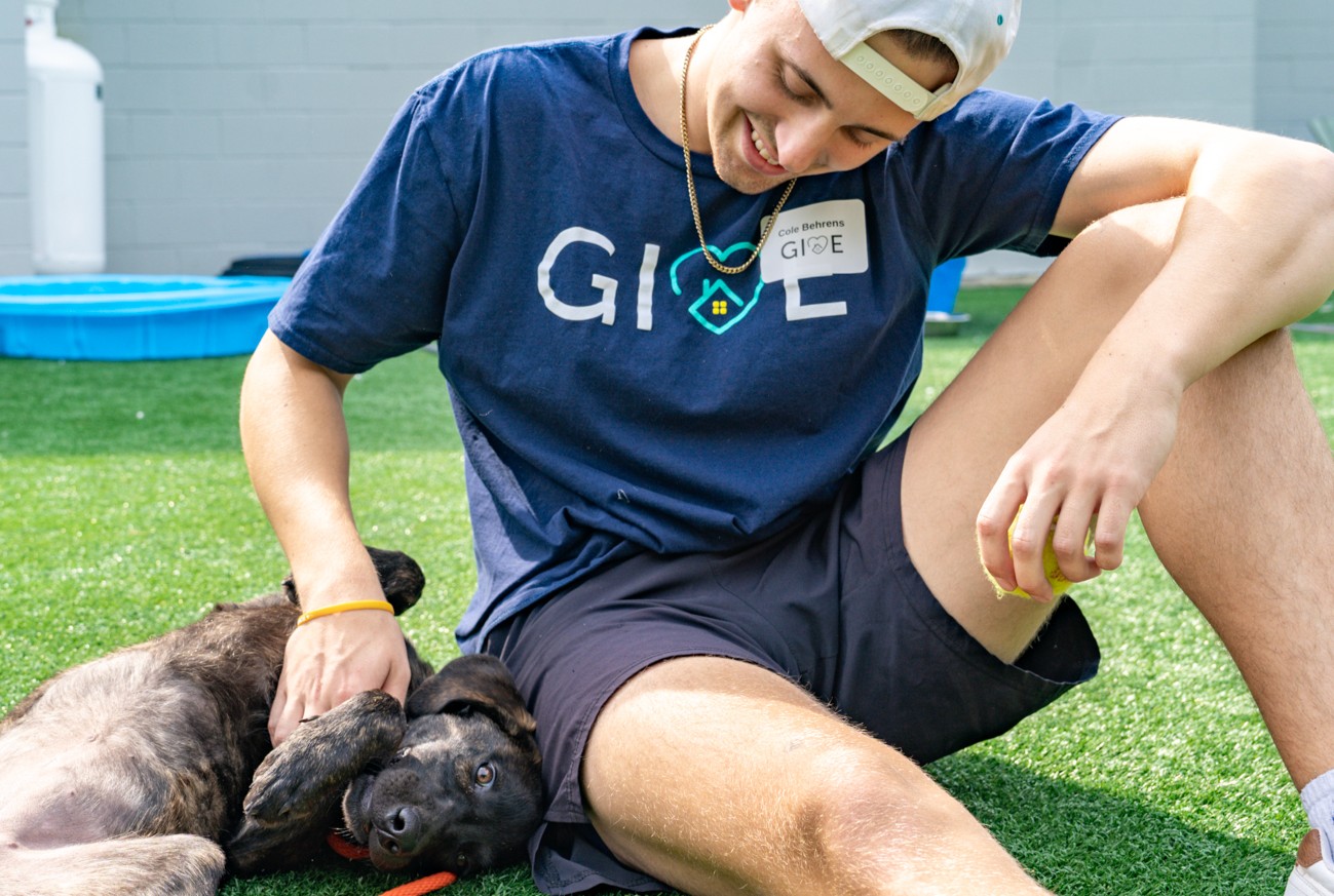 A young man in a navy t-shirt and shorts sits on grass, smiling while petting a playful black puppy lying on its back beside him. The man wears a backward cap and a name tag, and there's a blue kiddie pool in the background.