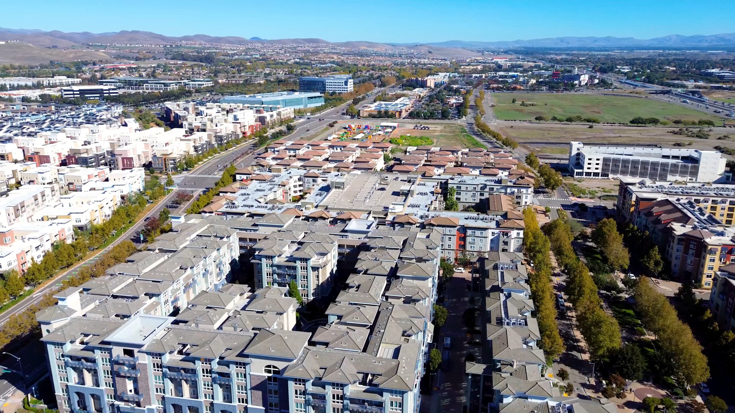 Aerial view of a suburban neighborhood with rows of houses, apartment buildings, streets, and green spaces, set against hills and an expansive cityscape under a clear blue sky.