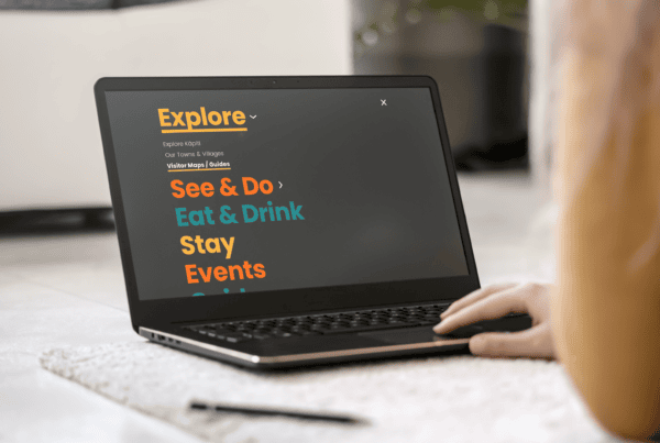 A person using a laptop displaying a Kāpiti regional tourism website section titled "Explore" with categories: See & Do, Eat & Drink, Stay, Events. The text is brightly colored on a dark screen. The scene includes a carpet and a plant in the background.