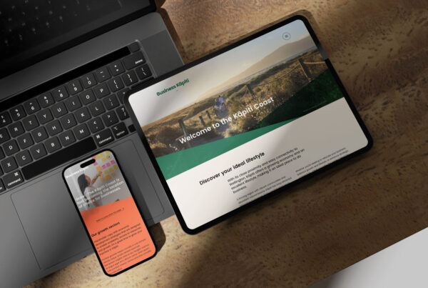 A tablet displaying a welcome screen for the Kapiti Coast Business website with a scenic image and text, placed on a wooden table next to a smartphone showing an orange and green app screen, and a partially visible laptop keyboard.
