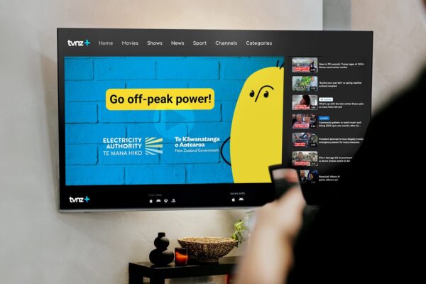A TV screen displays a TVNZ+ streaming app with a blue and yellow ad saying "Go off-peak power!" from the Electricity Authority of New Zealand. A person holds a remote, preparing to select content.