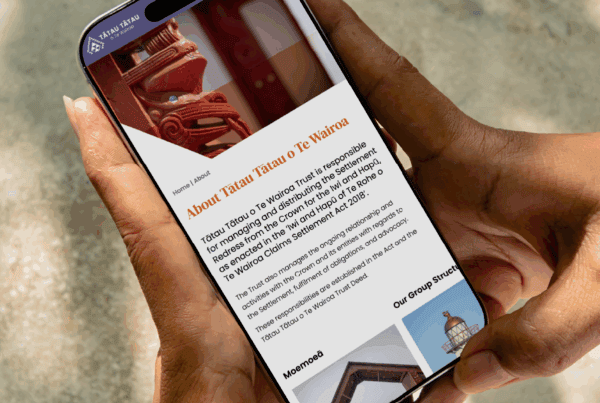 A person holds a smartphone displaying a webpage titled "About Tatau Tatau o Te Wairoa," featuring text and an image of a carved wooden figure, with sunlight illuminating their hands.