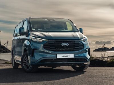 Ford Transit Custom 2024+ i blå, set forfra mod havet og skyerne.
