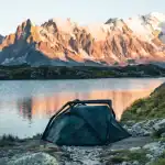Camouflage tent set up near mountain lake with scenic mountain backdrop.
