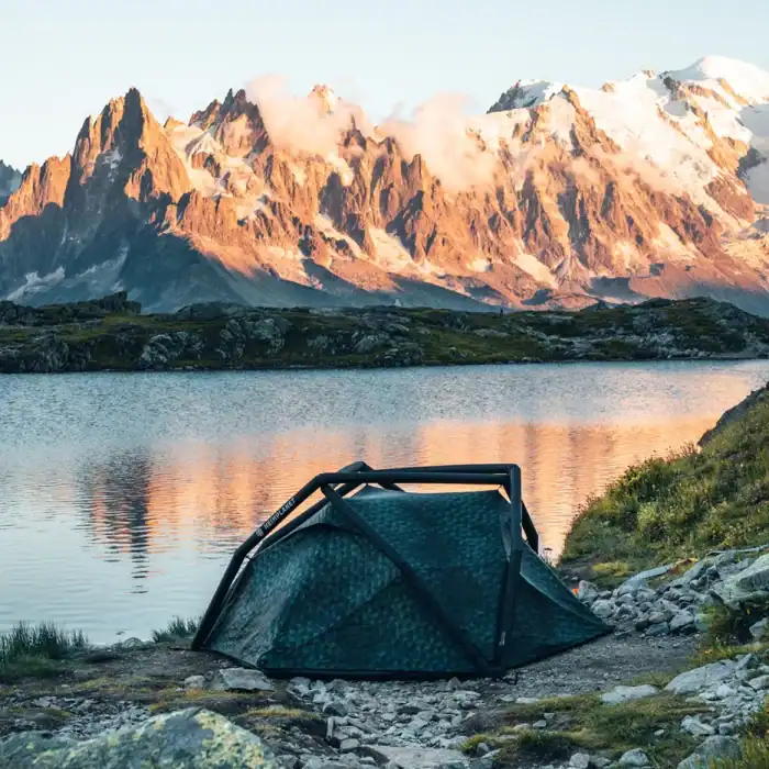 Camouflage tent set up near mountain lake with scenic mountain backdrop.