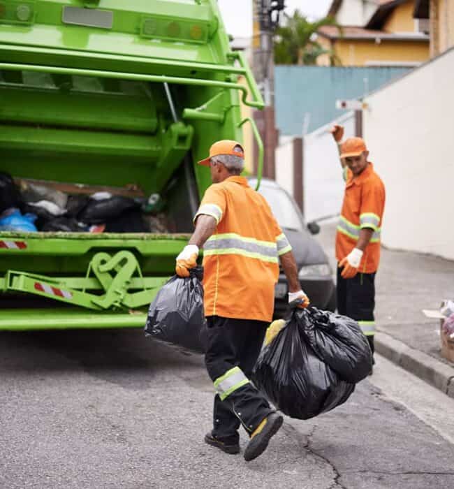 Garbage collection workers loading trash bags into a green dumpster for waste disposal.