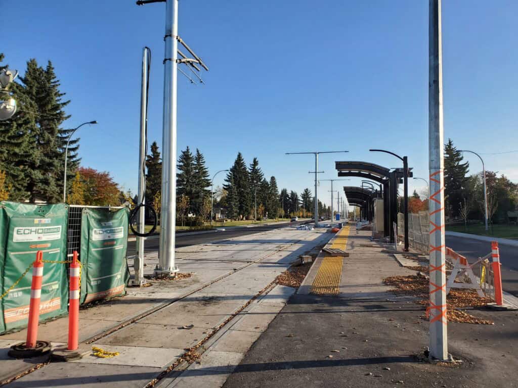 Uneven construction of tram stop with fencing, shelter, and electrical poles on a sunny day in a suburban area with trees.