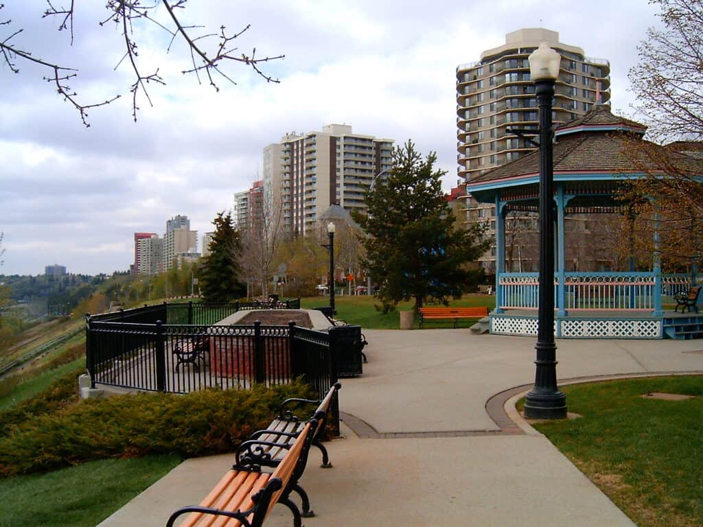 Colorful park gazebo with city high-rise buildings, trees, benches, and lampposts in an urban landscape during daytime.