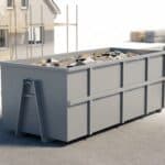 A large gray waste container filled with construction debris at an active building site, with stacks of wood and concrete structures in the background.