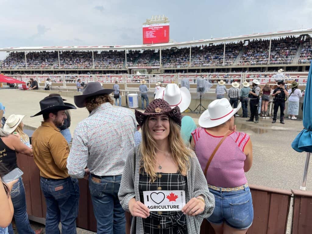 Young woman at Canadian rodeo wearing cowboy hat, holding "I love agriculture" sign, surrounded by rodeo fans in western attire.