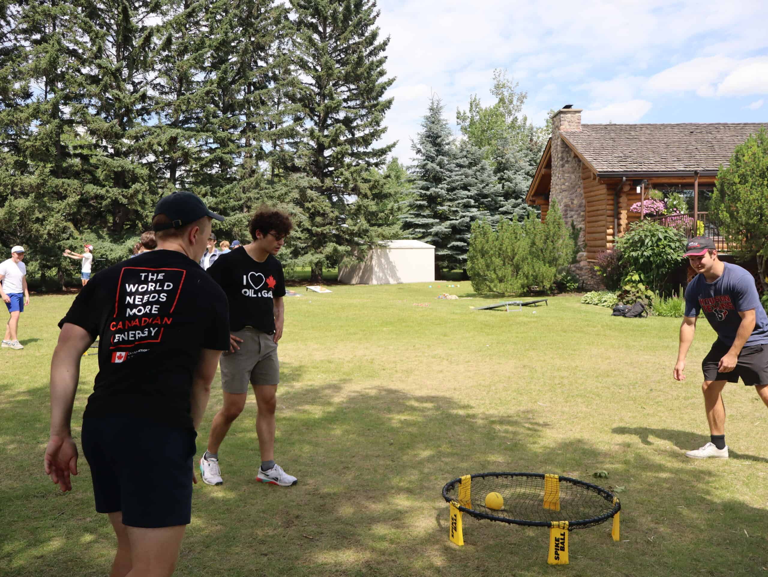 Grass field with people playing spikeball outdoors on sunny day, young Canadians for resources youth event promoting Canadian energy, sustainability, and resource development, natural landscape with trees and house in background.