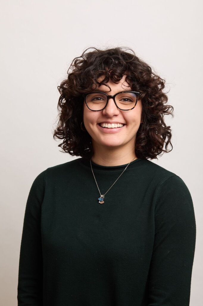 Young woman with curly brown hair, glasses, and a black sweater smiling against a plain background, representing young Canadians engaged in resource sector advocacy.
