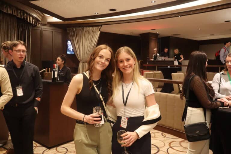 Two women smiling at a networking event, holding drinks, with a conference setting in the background.