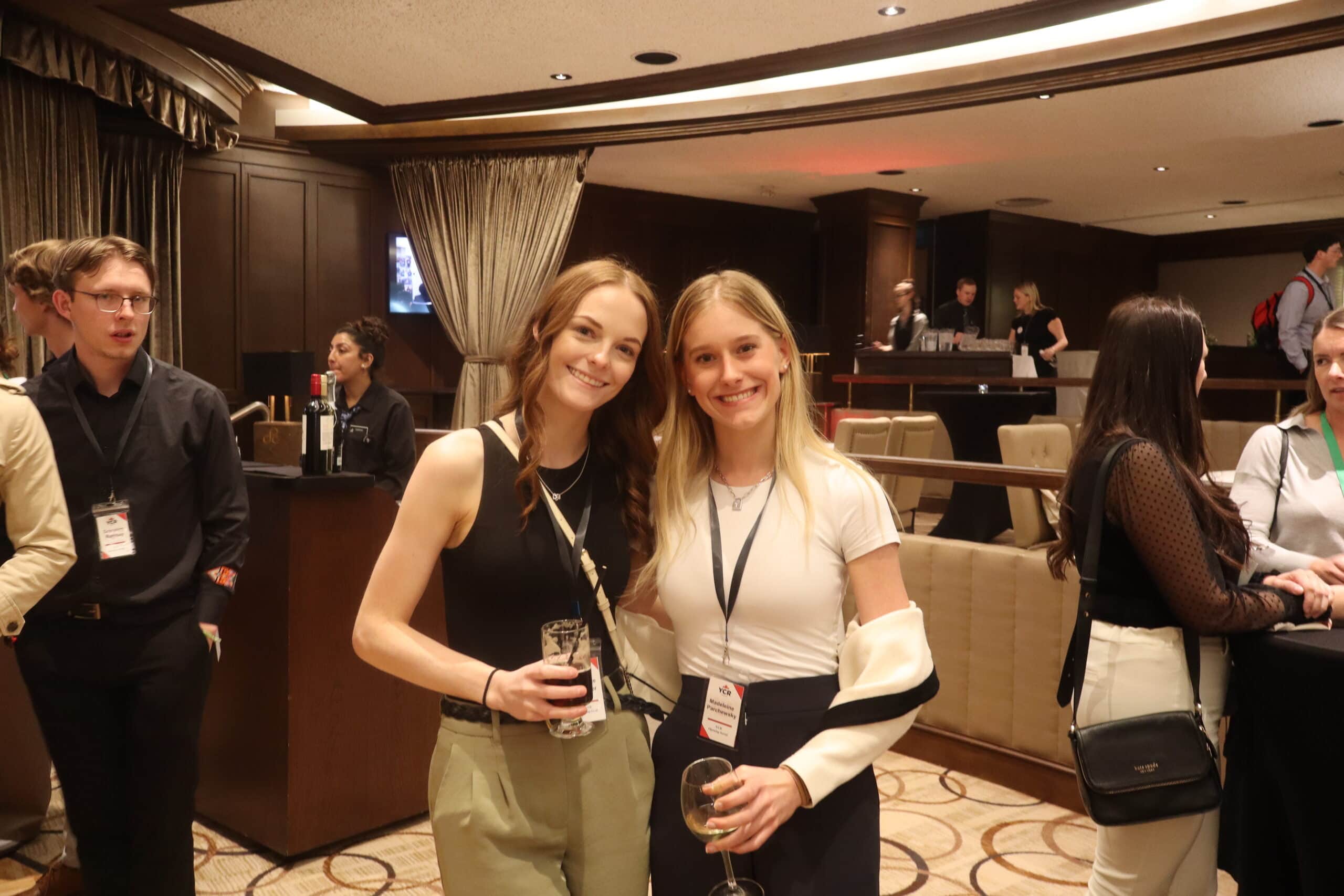 Two women smiling at a networking event, holding drinks, with a conference setting in the background.