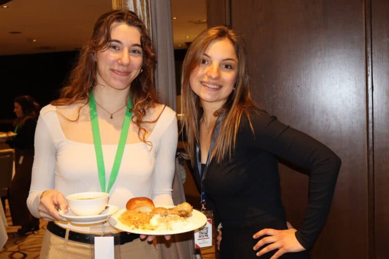 Two women smiling at a conference, holding a tray with coffee and pastries, in a professional setting.
