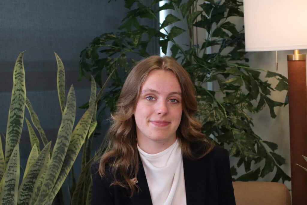 Portrait of a young woman with wavy brown hair, wearing a black blazer and white top, sitting in a modern office with green plants and a lamp.