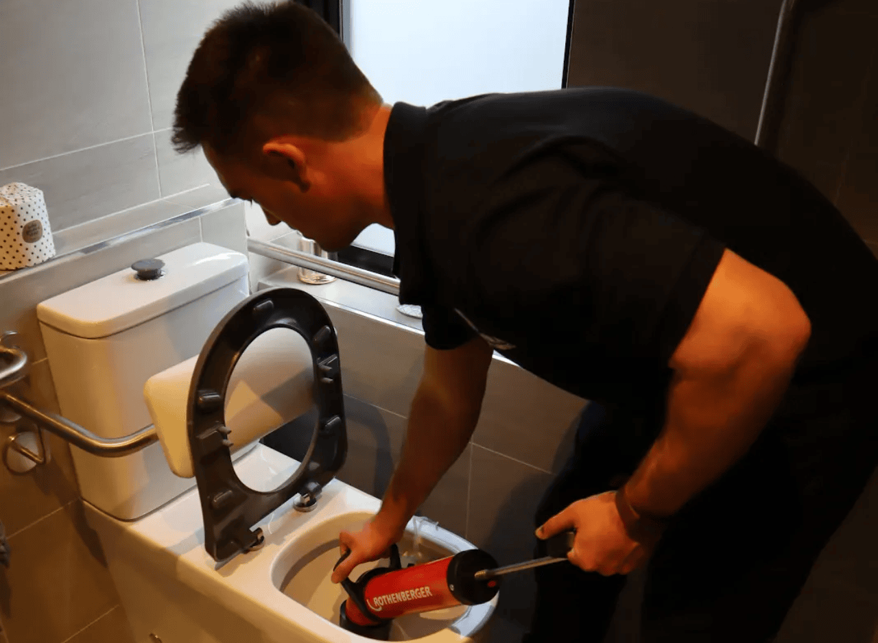 A plumber uses a drain cleaning tool to unclog a toilet in a modern bathroom with grey tiles and an elevated toilet seat.