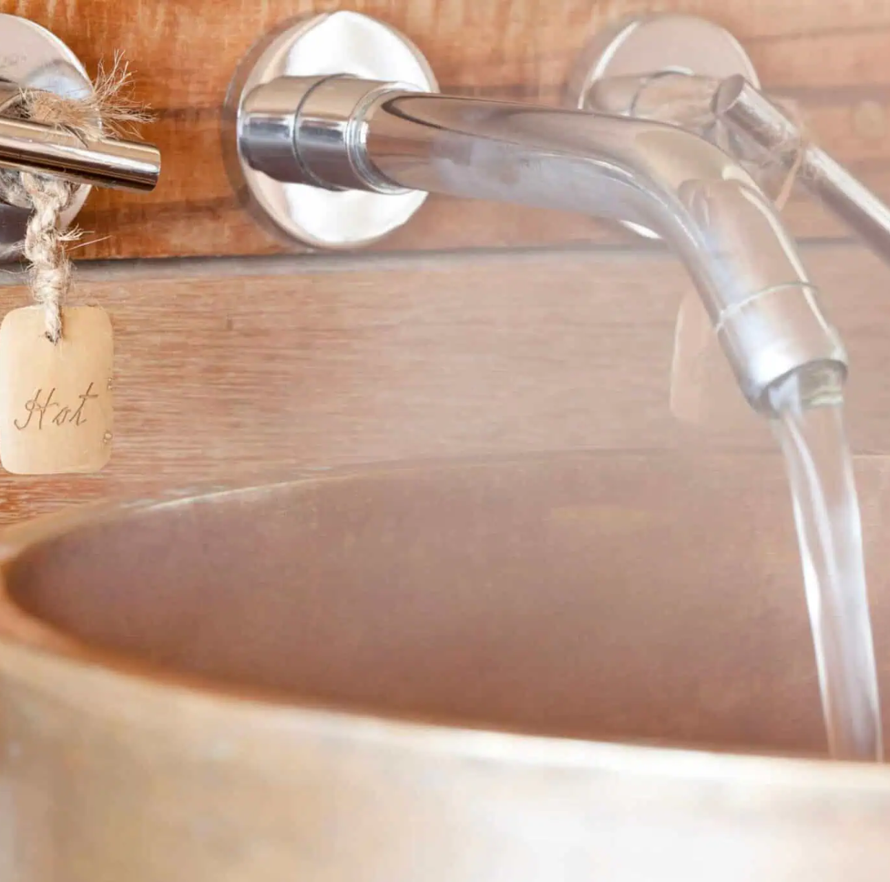 Hot water flows from a chrome wall-mounted tap into a rustic basin, with steam rising.