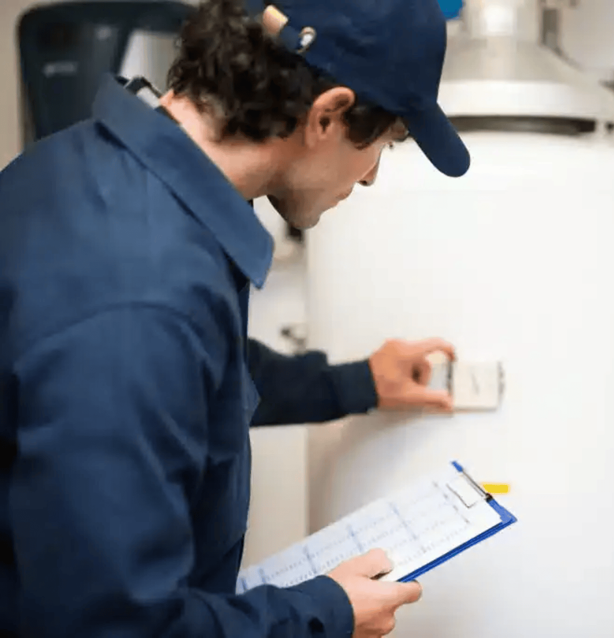 A technician in uniform checks the settings on a hot water heater while holding a clipboard for inspection notes.