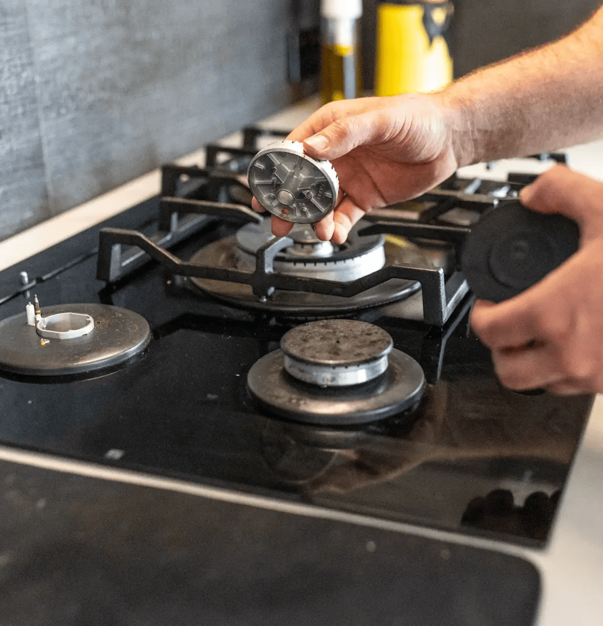 A close-up of a plumber's hands inspecting and replacing gas stove burner components on a black stovetop.