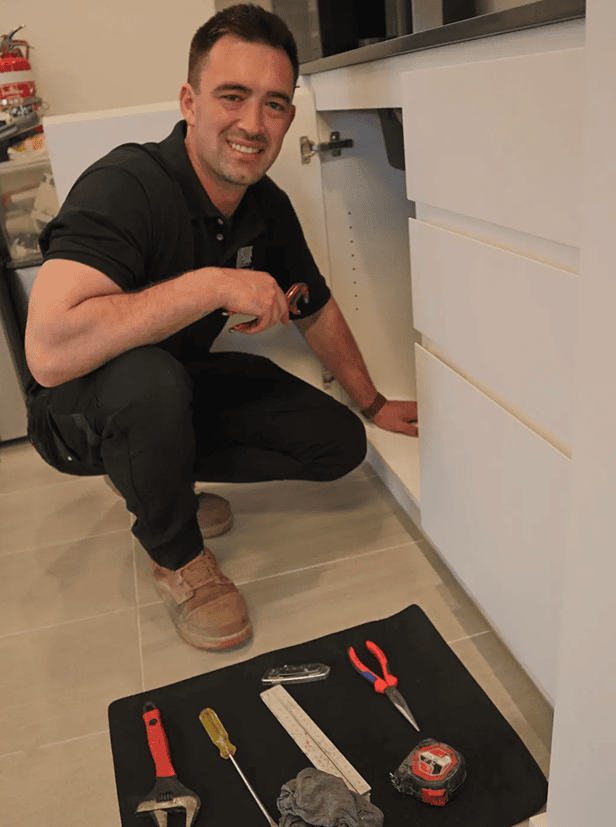 Smiling plumber inspecting plumbing under a kitchen sink during a service call.