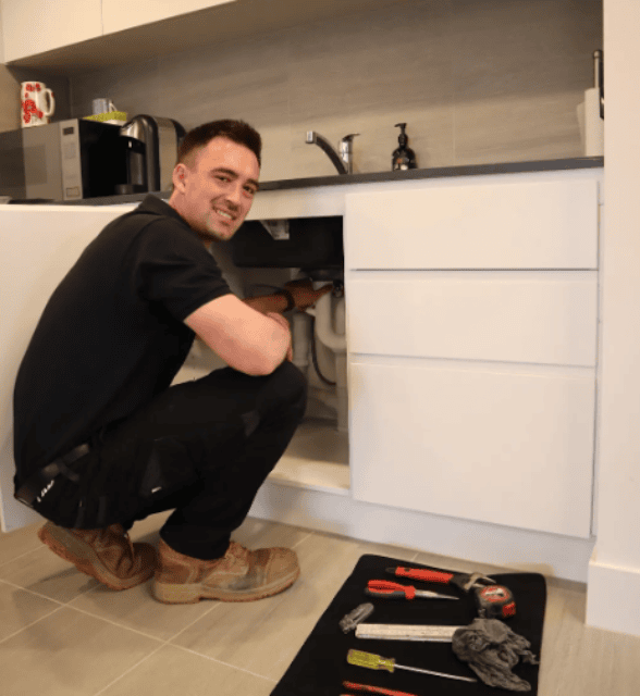 Friendly plumber crouched under kitchen sink, ready to perform maintenance with tools laid out.