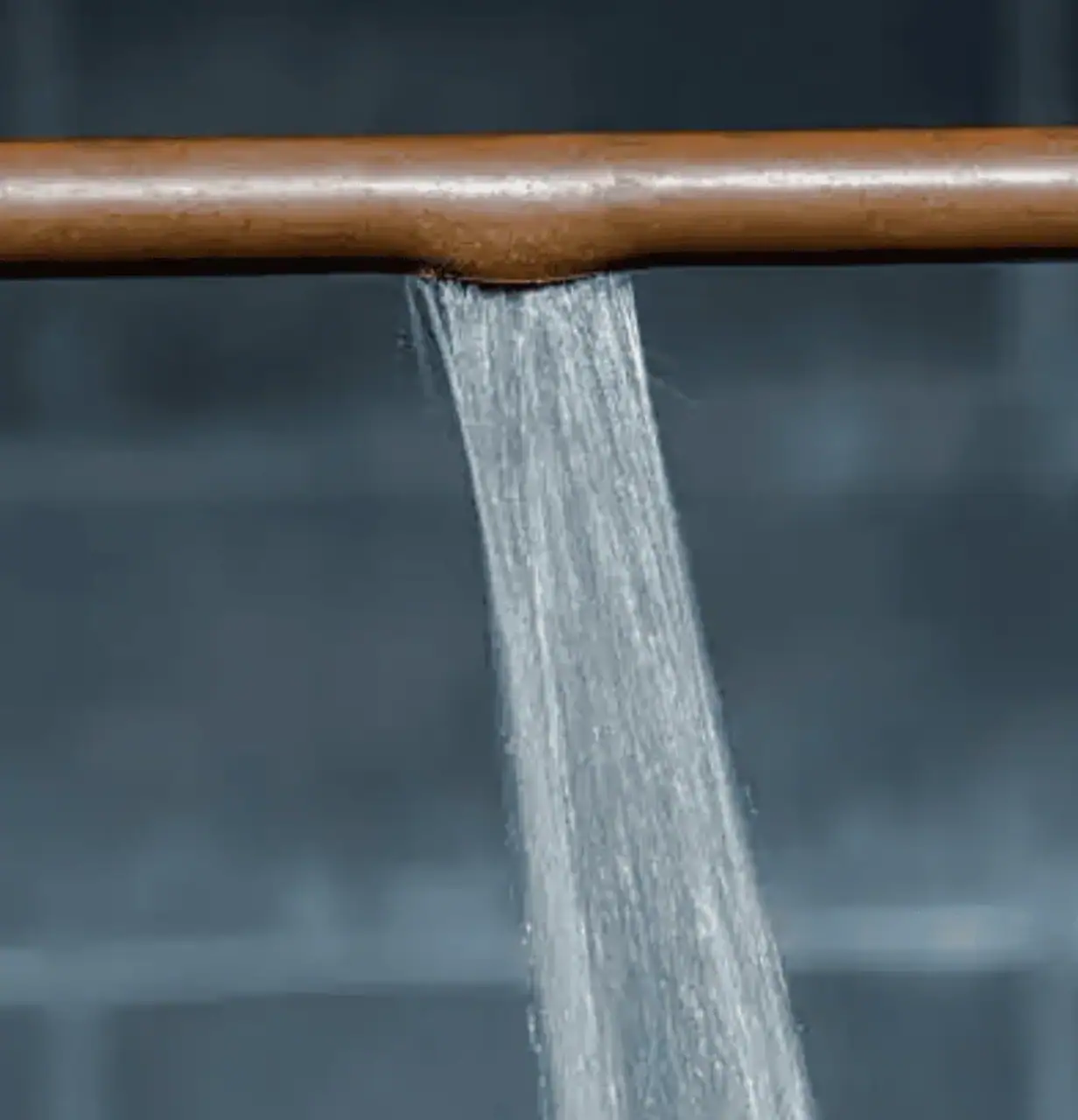Close-up of a copper pipe with a high-pressure water leak spraying a steady stream downward against a blurred background.