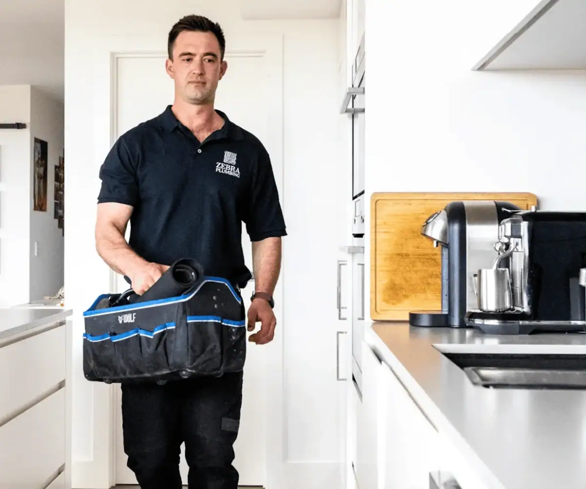 A plumber in a black uniform walks through a modern white kitchen carrying a blue and black tool bag and a rolled-up mat.