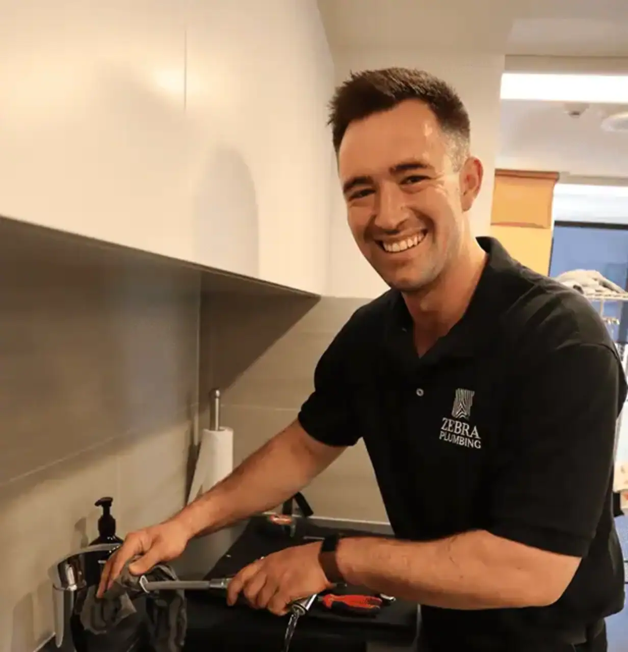 A smiling plumber in a black Zebra Plumbing polo cleans a modern kitchen faucet over a dark sink with a grey cloth.