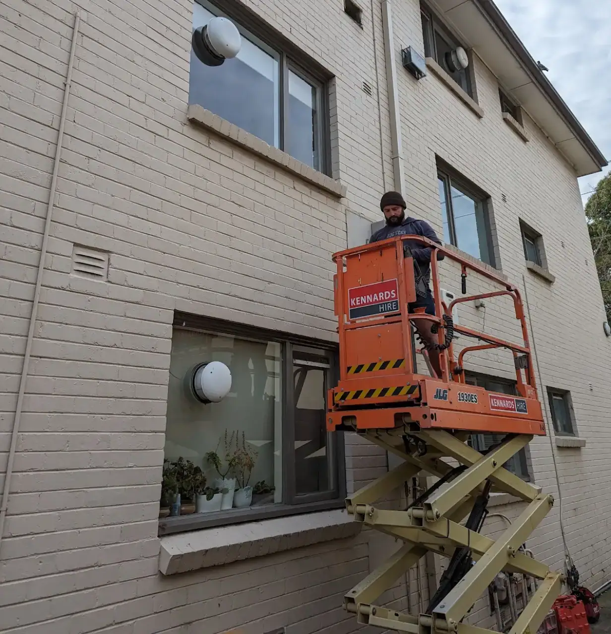 A worker on an orange scissor lift installing new gas hot water on the exterior of a three-story tan brick apartment building.