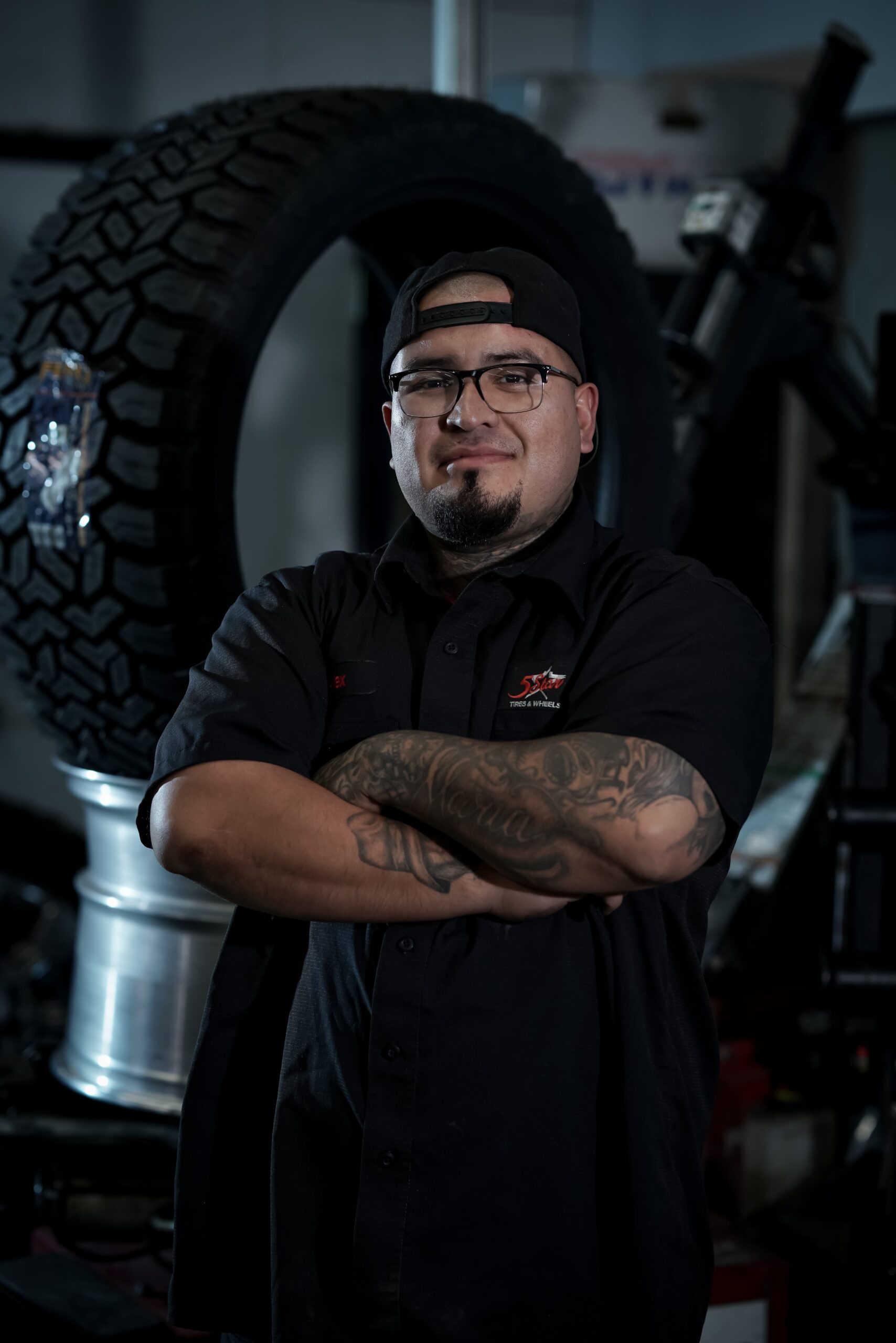 Confident tire technician with arms crossed in front of large tire at 5 Star Tires and Wheels.