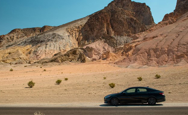 Black sedan driving through desert landscape with colorful rocky hills in the background.