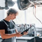 Auto repair technician inspecting vehicle undercarriage with diagnostic tablet.