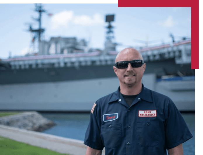 Skilled Army mechanic in uniform with naval ship in background.