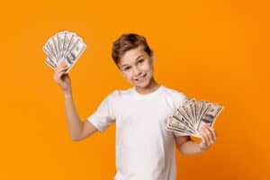 Cute teen boy holding fans of money and smiling, orange studio background.