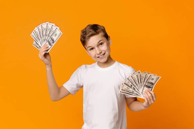 Cute teen boy holding fans of money and smiling, orange studio background.