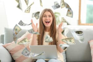 Surprised young woman with laptop and flying money at home