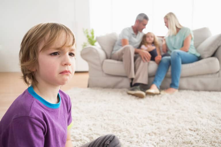 Upset boy sitting on floor while parents enjoying with sister on sofa at home